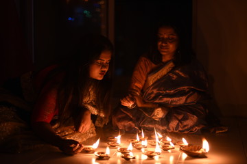 Two beautiful Indian Bengali women in Indian traditional dress are lighting Diwali diya/lamps sitting on the floor indoor in darkness on Diwali evening. Indian lifestyle and Diwali celebration