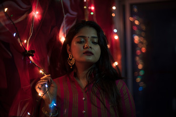 Portrait of an Indian Bengali beautiful brunette woman in front of the colorful light bokeh background created by tiny chain lights in the evening of Diwali. Indian lifestyle and religion.