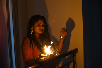 Two young and beautiful Indian Bengali women in Indian traditional dress are celebrating Diwali with diya/lamp and fire crackers on a balcony in darkness. Indian lifestyle and Diwali celebration