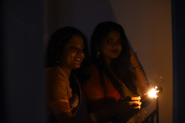 Two young and beautiful Indian Bengali women in Indian traditional dress are celebrating Diwali with diya/lamp and fire crackers on a balcony in darkness. Indian lifestyle and Diwali celebration