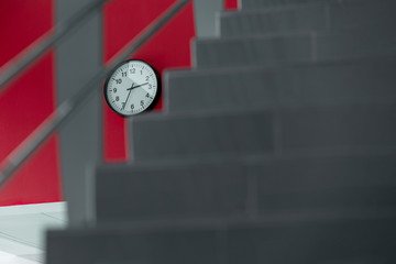 Obraz premium A round white clock is on the wall on a geometric red and gray background and a blurred stairs. Time management concept.