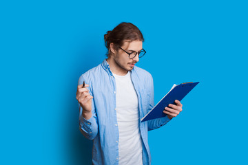 Caucasian bearded boy with long hair is looking in a notebook through eyeglasses and posing on a blue wall