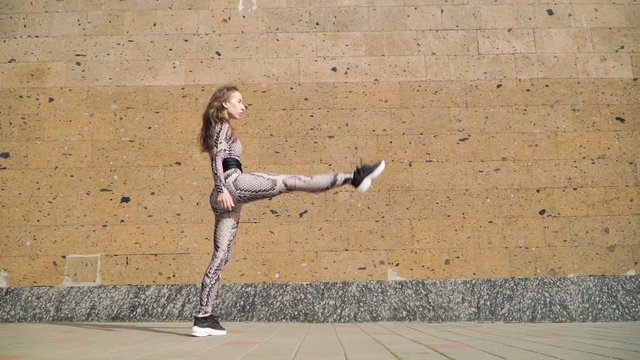 Young Beautiful Athletic Girl Gymnast with Steep Stretch. She goes and does leg in split. Dressed in her slinky jumpsuit with picture scales Cobra. On a brown background concrete wall. Slow Motion