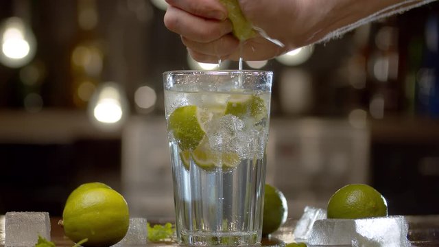 Close up male hand squeezing lime into a glass with tonic, soda water or cocktail. Glass with a refreshing drink, ice, limes and leaves on a table against the blurry lights background