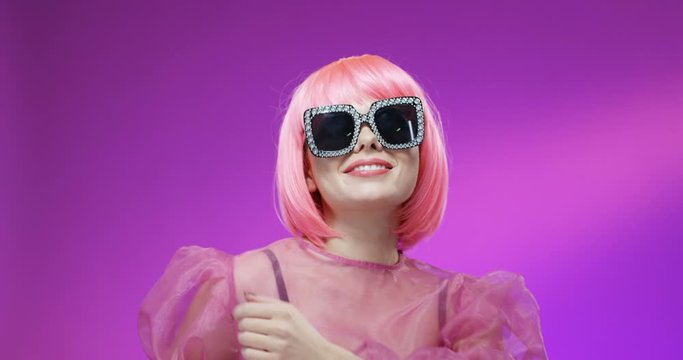 Portrait of Caucasian joyful stylish woman in pink wig and glamourous sunglasses smiling to camera on velvet background and blowing kisses. Close up of happy girl dancing. Extraordinary doll female.