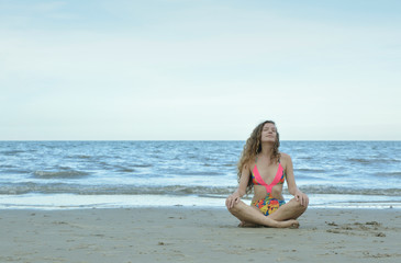 Pretty beautiful woman in pink bikini meditating and sitting on the beach over sea and sky, Healthy and fitness concept