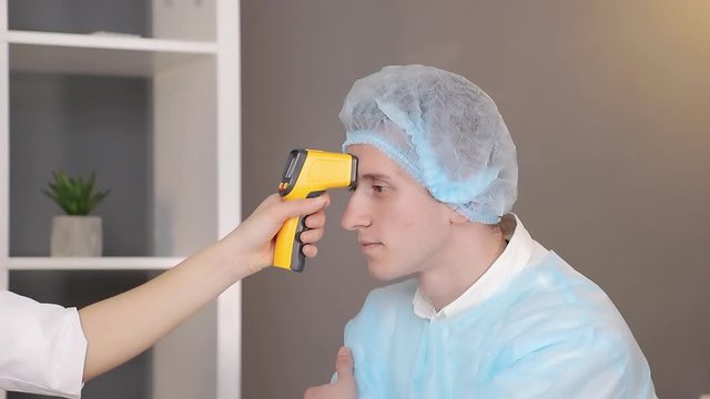 Doctor in a white coat and a medical mask measures the temperature of a patient in the medical office with an infrared non-contact thermometer.