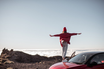 Young woman dressed in red enjoying rocky landscapes above the clouds, standing on the car highly...