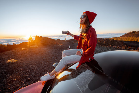 Young woman dressed in red enjoying sunset landscape above the clouds, sitting on the car roof while travel in the mountains. Carefree lifestyle and travel concept