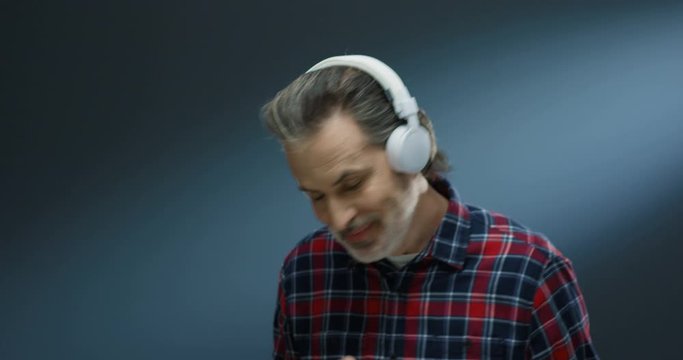 Caucasian cheerful senior man with gray hair in big headphones listening to music and dancing funny on grey wall backgrund. Male imitating playing on guitar and pretending musician.