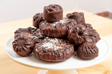 Delicious chocolate dietary candies and cookies for vegans made from natural ingredients. Tasty treats, sprinkled with coconut, on a white tray on wooden table. Healthy meal. Sweet and harmless.