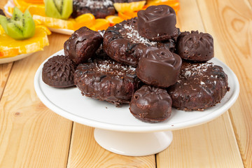Tasty treats, sprinkled with coconut, on a white tray on wooden table. Delicious chocolate dietary candies and cookies for vegans made from natural ingredients. Healthy meal. Sliced fruits in back.