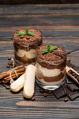 Classic tiramisu dessert in a glass cup, pieces of chocolate and savoiardi cookies on wooden background