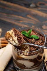 Classic tiramisu dessert in a glass cup, pieces of chocolate and savoiardi cookies on wooden background