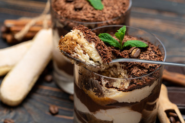 Classic tiramisu dessert in a glass cup, pieces of chocolate and savoiardi cookies on wooden background