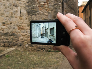 A hand holding a camera with a picture on the screen: an outdoor photographer taking a picture of a young couple standing in the ancient street