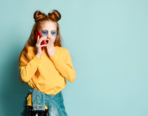 Surprised red-haired teen girl in blue smoked glasses and yellow sweatshirt is talking by smartphone