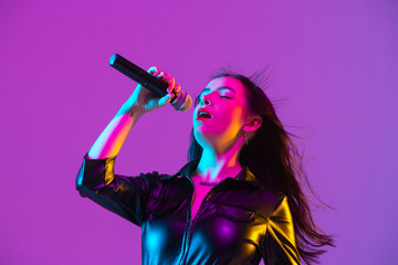 Caucasian female singer portrait isolated on purple studio background in neon light. Beautiful female model in black wear with microphone. Concept of human emotions, facial expression, ad, music, art.