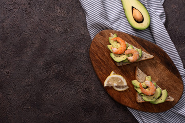 Wholegrain toast bread slices with guacamole, fried shrimp and fried bacon pieces on wooden board Selective Focus, Focus on the front of the shrimp on the first bread