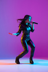 Obraz premium Caucasian female singer portrait isolated on purple studio background in neon light. Beautiful female model in black wear with microphone. Concept of human emotions, facial expression, ad, music, art.