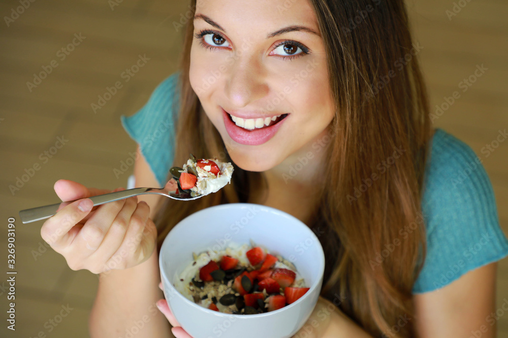 Wall mural close up from above of beautiful young woman smiling and eating skyr with cereal muesli fruit and se