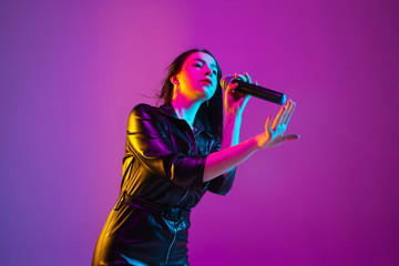 Caucasian female singer portrait isolated on purple studio background in neon light. Beautiful female model in black wear with microphone. Concept of human emotions, facial expression, ad, music, art.