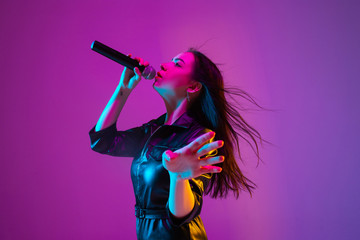 Caucasian female singer portrait isolated on purple studio background in neon light. Beautiful female model in black wear with microphone. Concept of human emotions, facial expression, ad, music, art.