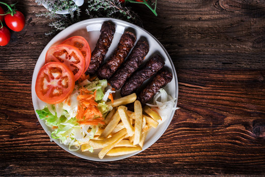 Spicy kofte (cevapcici, grilled minced meat) served with fresh tomatoes, lettuce, salad, vegetables and French fries on a rustic wooden board