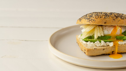 Avocado, soft cheese, poached egg sandwich lying on a round plate on a right side. Egg yolk flow out. Wooden white background. 
