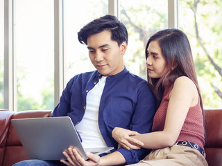 Young Asian couple sitting close together  on couch in living room , a man using computer while woman holding his arm , smiling and looking at his face