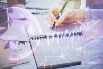 Hand taking notes in notepad. Forex chart holograms in front. Concept of research. Double exposure