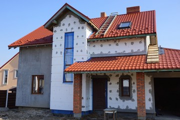 large unfinished private house with white insulation on a gray wall under a red tile roof against a blue sky on a sunny day