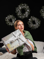 A beautiful girl with blond hair and green eyes in light clothes is sitting on a bed and is holding a wrapped gift in her hands