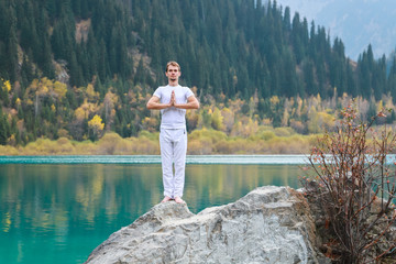 A young man in white practices yoga in the mountains lake. Pose Samasthiti namaskar