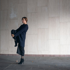 Woman in a coat and high heels stands on one leg in an urban environment