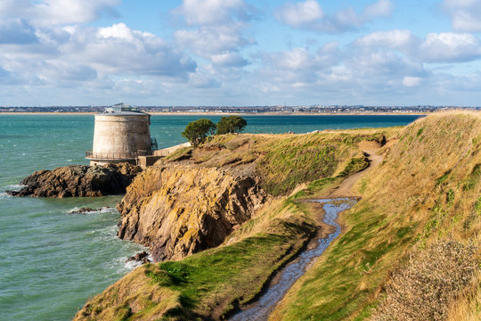 Iconic Martello tower landmark in Sutton, Dublin, Ireland, view from the Howth Cliff Walk hiking path. Scenic Irish coastal trail next to the sea and sheer cliffs.