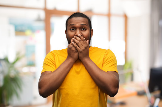 emotion, expression and people concept - scared young african american man in yellow t-shirt covering his mouth by hands over office background - Powered by Adobe