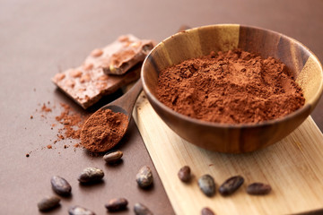 sweets, confectionery and culinary concept - chocolate with hazelnuts, cocoa beans and powder in wooden bowl with spoon on brown background