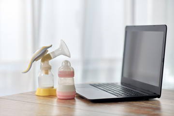 feeding and technology concept - bottle with baby milk formula, breast pump and laptop computer on wooden table at home