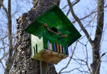 Green wooden birdhouse with  color pencils. It hanging on the tree in forest on sky background. Early spring, Russia