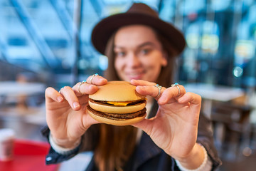 Happy stylish hipster woman eats burger bun with beef in fast food restaurant