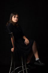 Fifteen-year-old girl sits on a bar chair, snasked on a black background in a dark