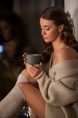 winter, comfort and people concept - young woman in pullover sitting at window with coffee or tea cup at home