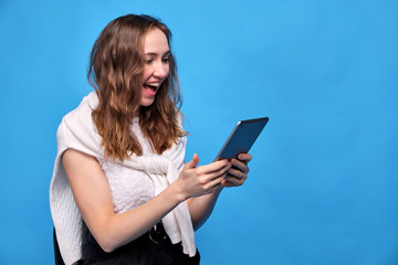 A girl in casual clothes on a blue background sits and looks at the tablet in surprise and rejoicing.