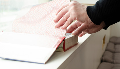 A person reading books near the window. Hands turns over book page.