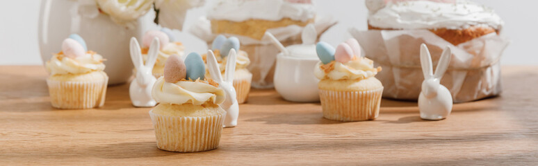 Selective focus of cupcakes with decorative bunnies, sugar bowl, easter cakes and vase on wooden background, panoramic shot