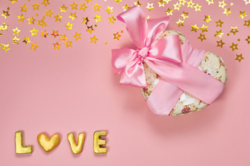Heart shaped Valentines Day gift box with pink ribbon, small gold stars and gold word love on paper background.