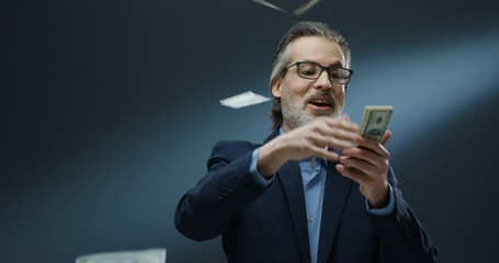 Cheerful rich handsome businessman of middle-age in glasses and suit tossing money on grey wall background. Happy good looking man in business style throwing dollars. - Powered by Adobe