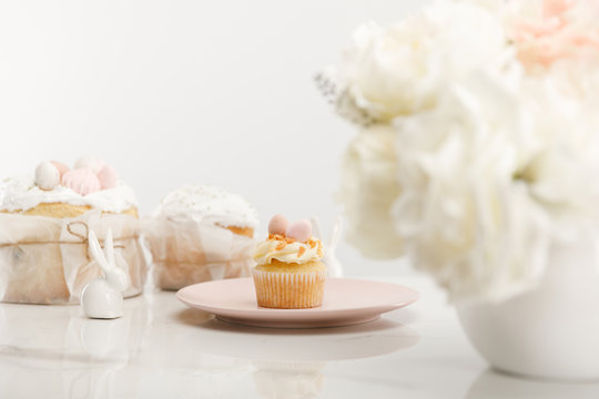 Selective focus of cupcake on plate, decorative rabbits, easter cakes and vase with bouquet on white background
