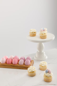 Delicious Easter cupcakes on surface and cake stand with painted chicken eggs on grey background